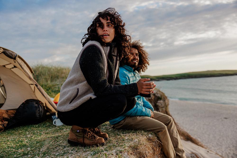 A woman in a black sweater, beige vest, and hiking boots sits on a rock overlooking a beach, with a tent and a man in a blue jacket in the background.