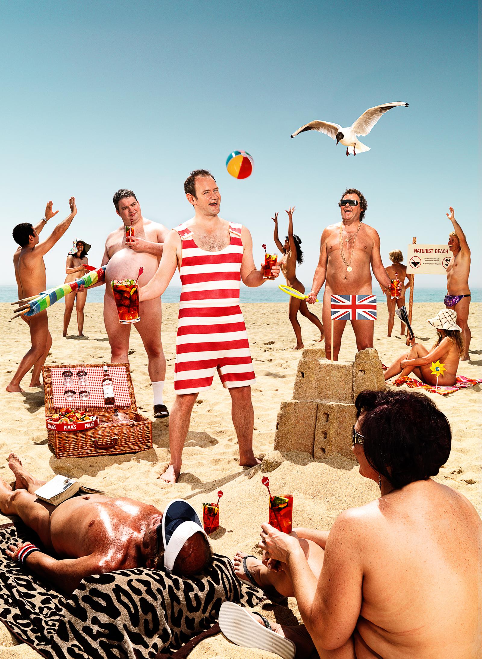 A group of people on a beach partying with a beach ball, picnic basket, and sandcastle, near signs reading 'Naturist Beach'.