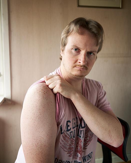 A man with a short, blond, messy hair, wearing a pink, sleeveless, graphic t-shirt, with his left hand on his right shoulder, in a dimly lit, beige, wall- and window-lined room.