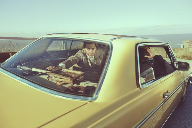A man in a suit and tie is driving a 1970s-style car, with a vintage typewriter and other retro office equipment visible in the back seat.