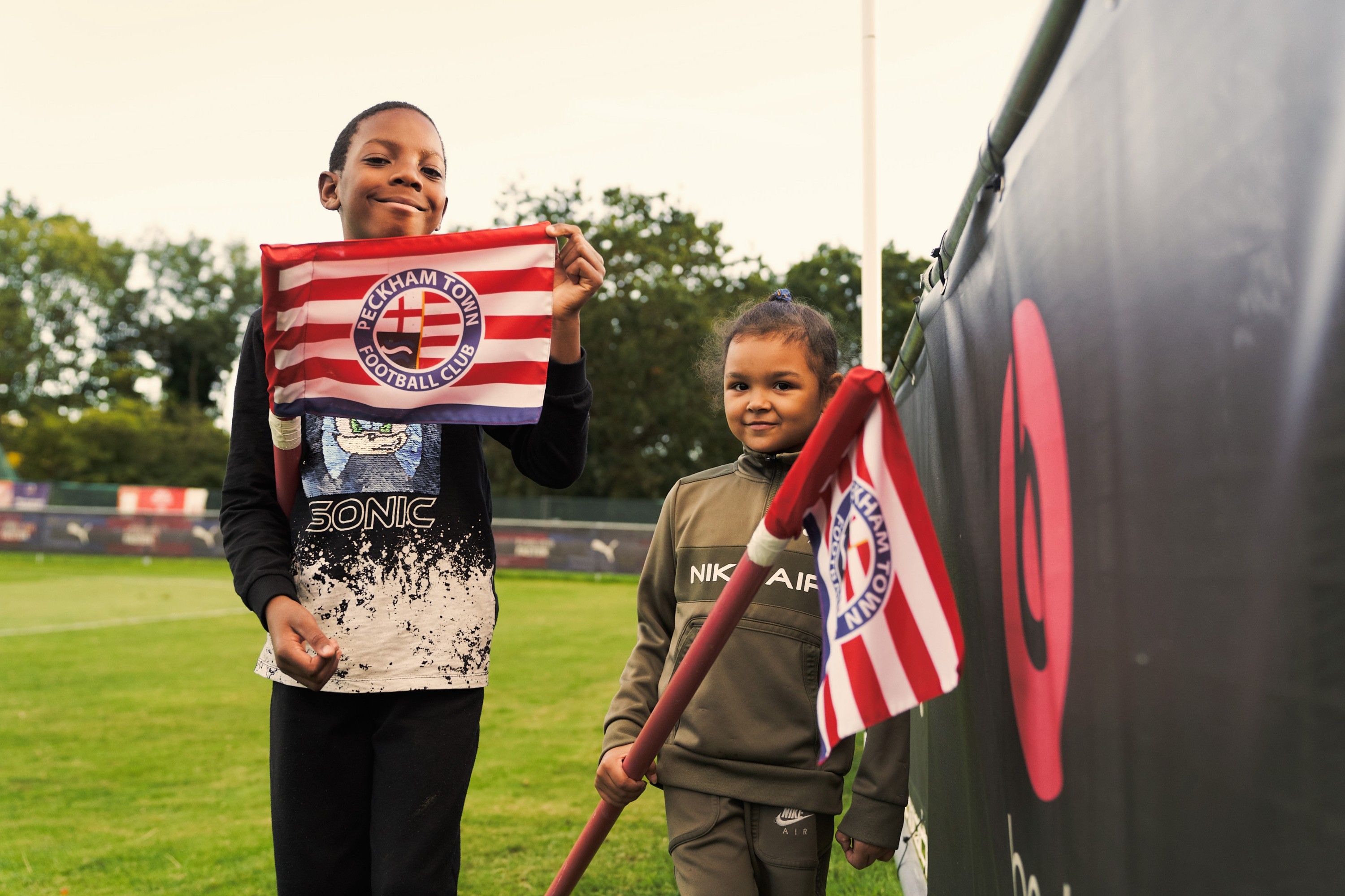 Two young children, one boy and one girl, holding a football club flag, standing on a football field.
