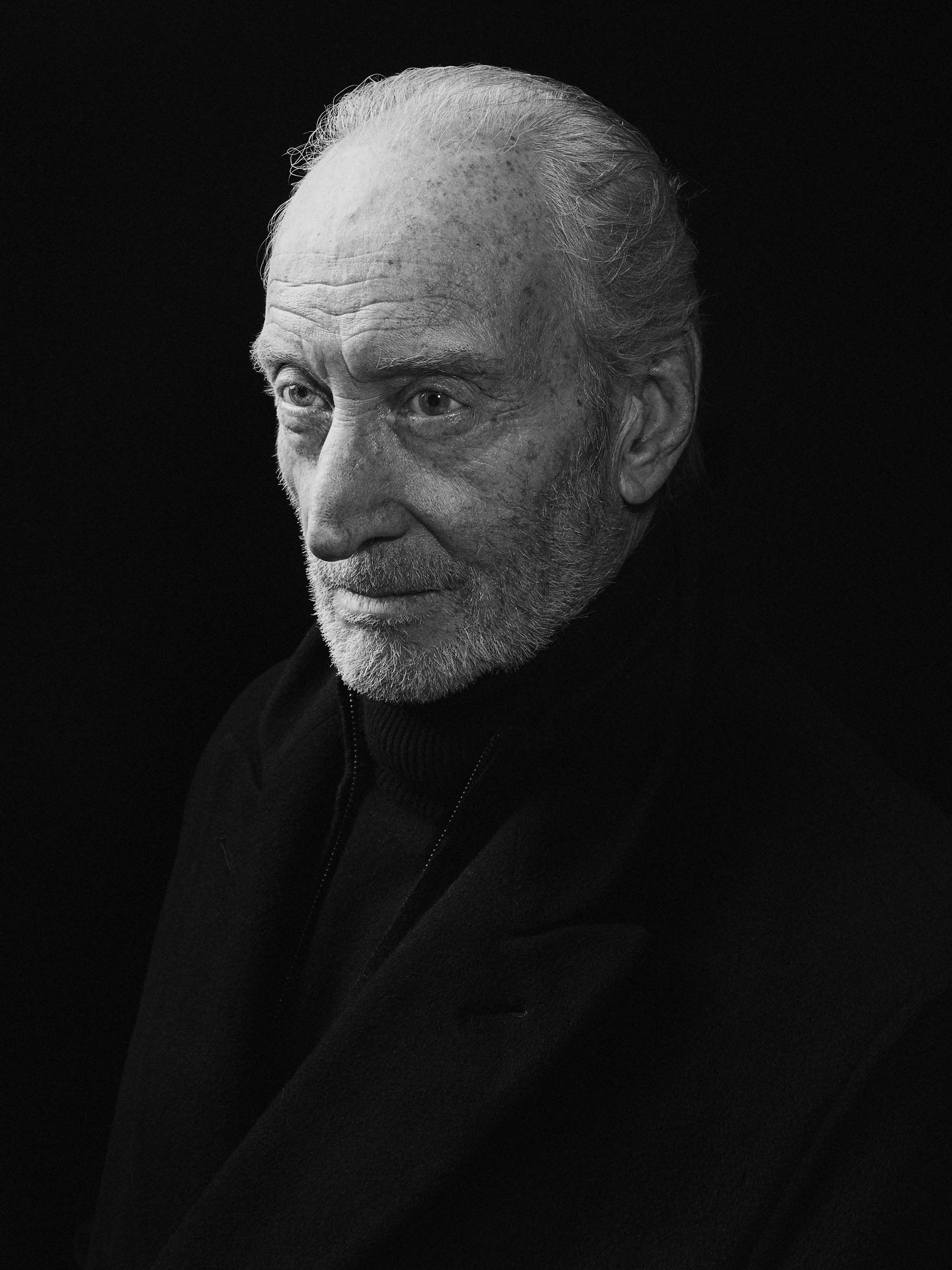A black and white portrait of an elderly man with a receding hairline, wearing a dark zip-up sweater.