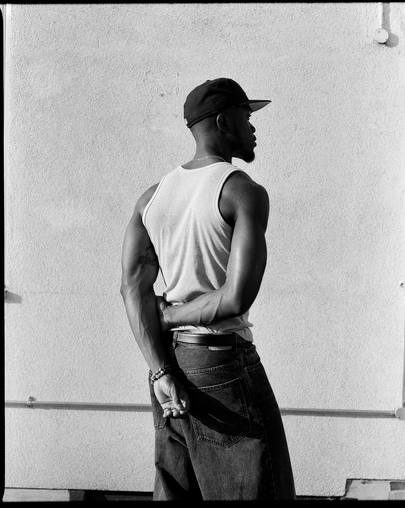 A man in a tank top and jeans, standing in front of a wall, looking to the side.