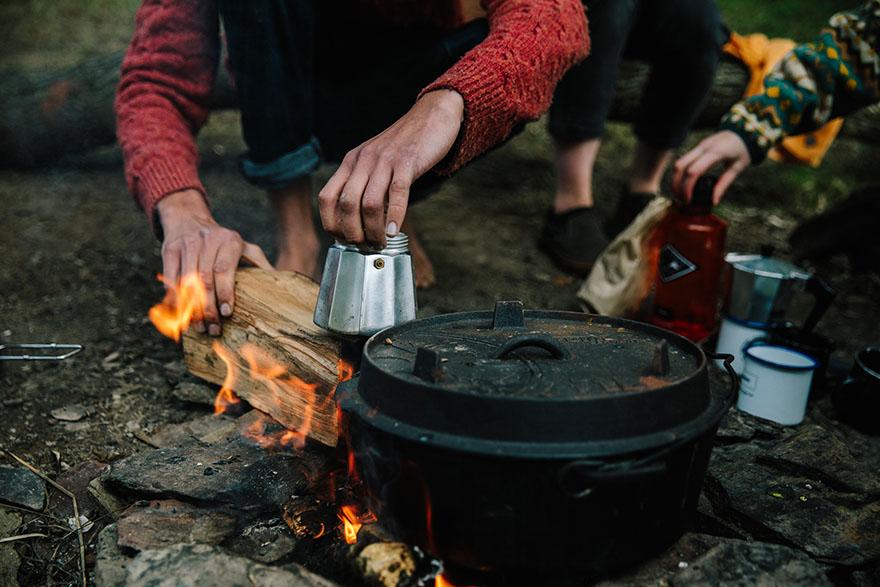 A person in a red sweater and jeans is gathering firewood near a campfire, with a pot and other camping equipment nearby.