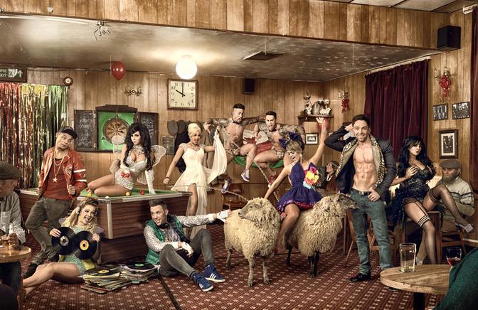 A group of people, including celebrities, in a pub-like setting, with a DJ spinning records, surrounded by a mix of people in various outfits, including a woman in a wedding dress, a man in a football jersey, and a person in a wetsuit, with a sheep in the background.