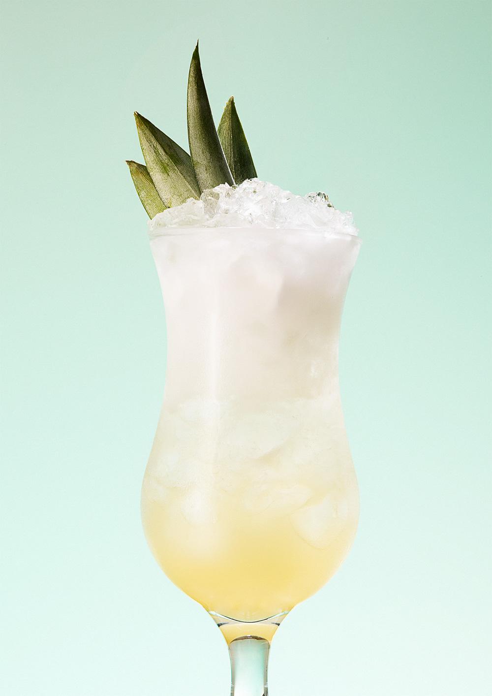 A tall glass containing a yellowish-white cocktail with a pineapple slice as a garnish.