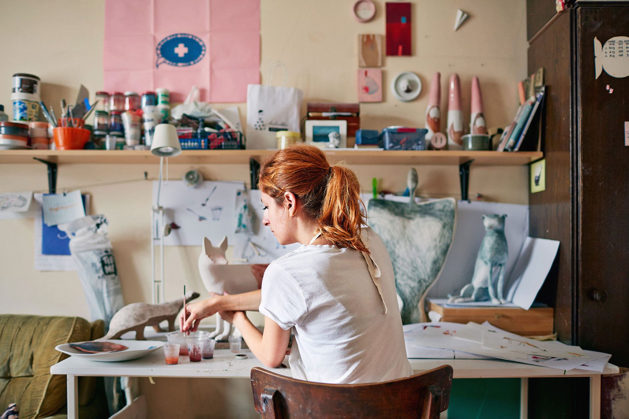 Artist Kerry Harrison sitting at a desk in a cluttered studio, surrounded by art supplies and half-finished sculptures, working on a new piece.