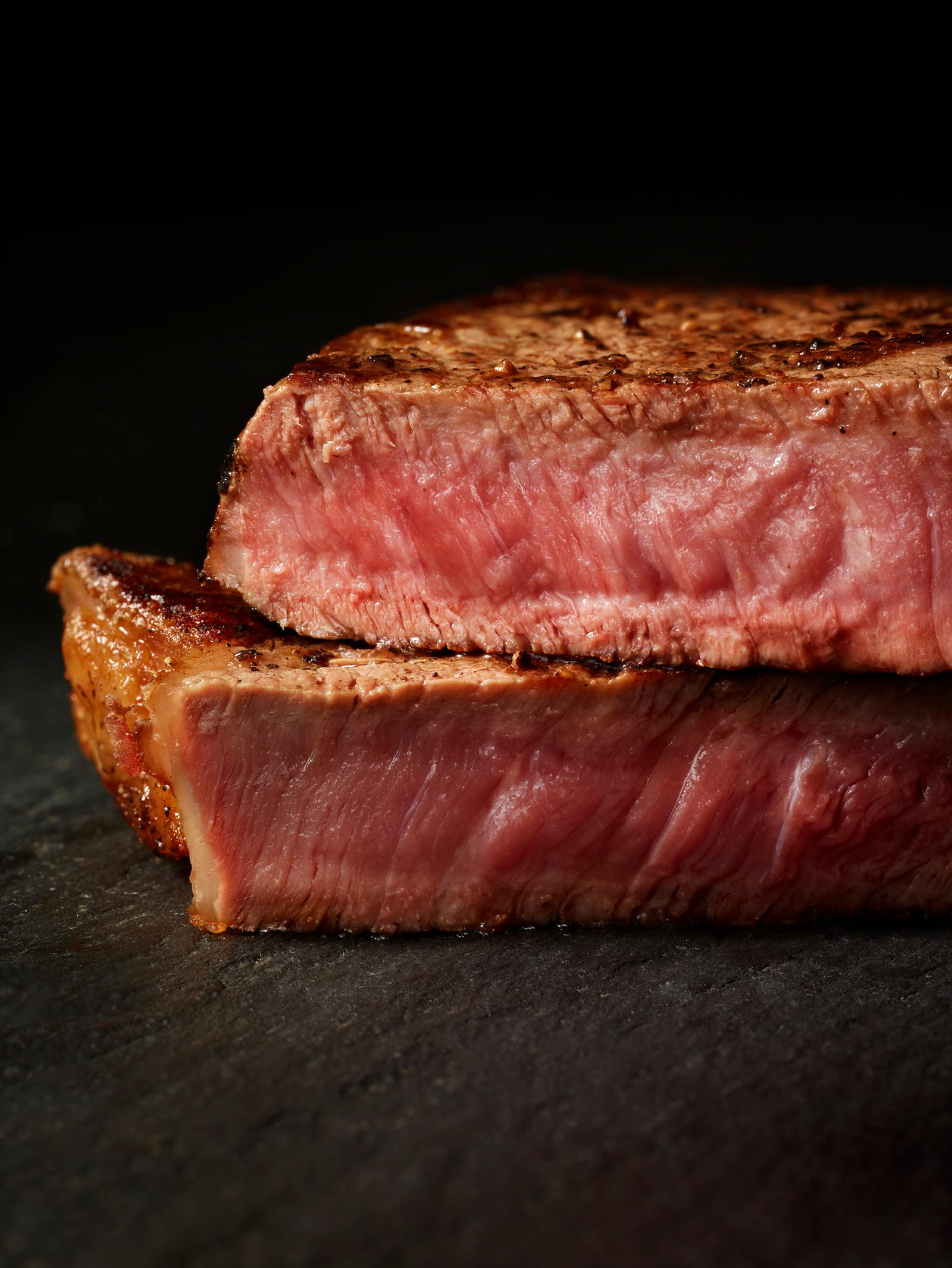 A cooked, sliced, and stacked piece of beef, likely a steak, with a pink, juicy, and slightly browned color, on a dark, possibly wooden, surface.