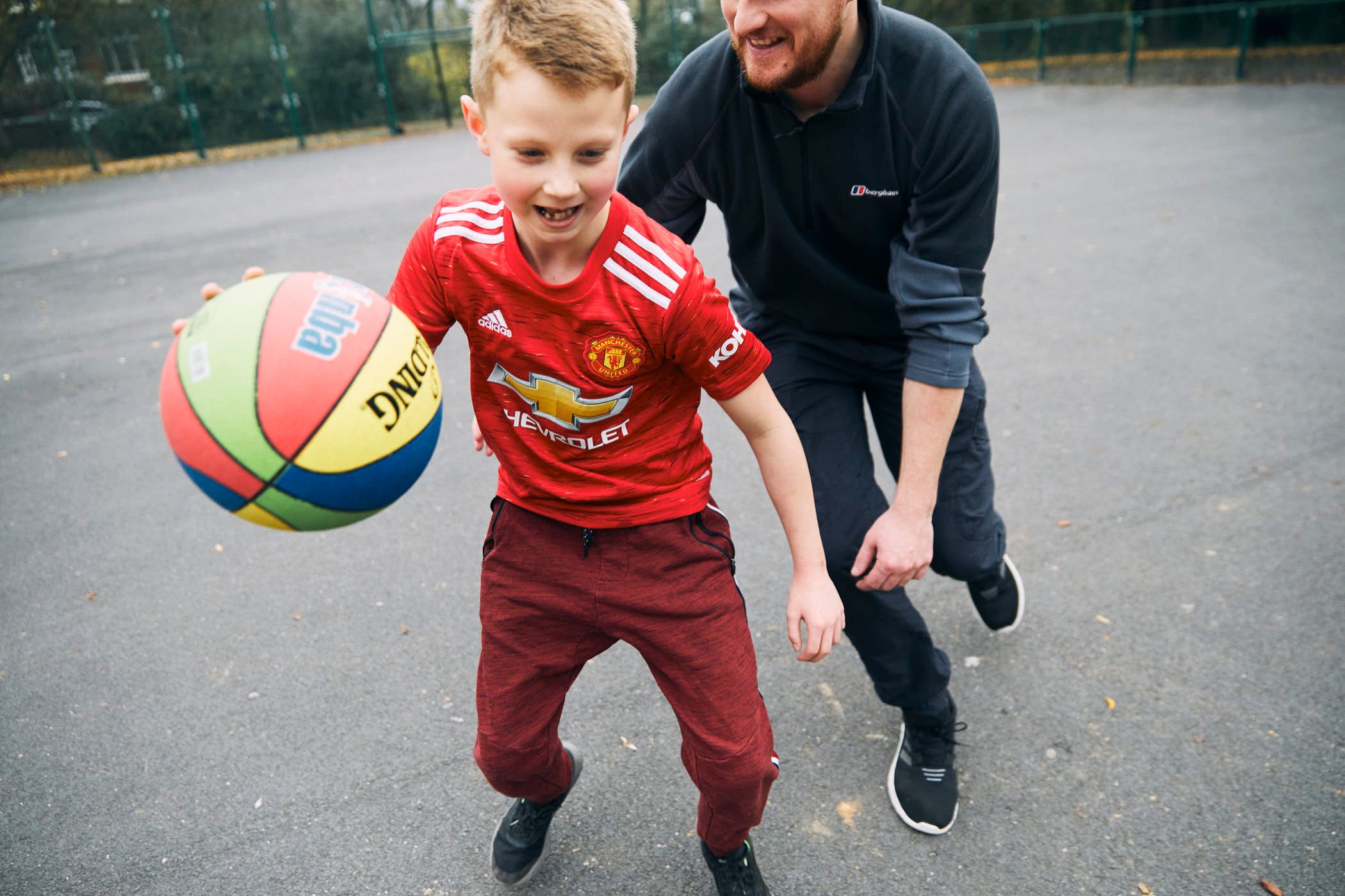 A young boy in a red Manchester United jersey is dribbling a ball with a man in a black polo shirt guiding him. The boy is smiling and the man is helping him with his movement.