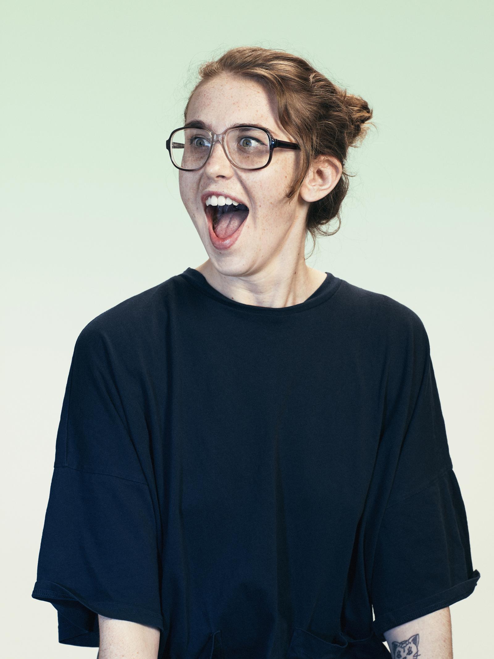 A woman with a shocked expression, wearing a black t-shirt and glasses, with her mouth open in a loud, exaggerated 'O' shape.
