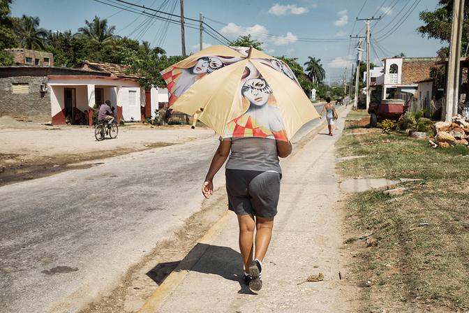 A woman walking down a street in a tropical location, wearing a yellow umbrella with a large, colorful, and cartoon-like image of a woman's face on it, with a red and yellow striped pattern on the umbrella's edges.