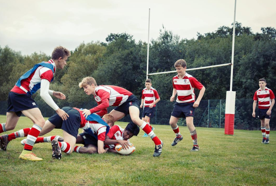 A group of young boys in rugby jerseys and shorts are playing rugby on a grassy field, with one boy lying on the ground and others surrounding him.