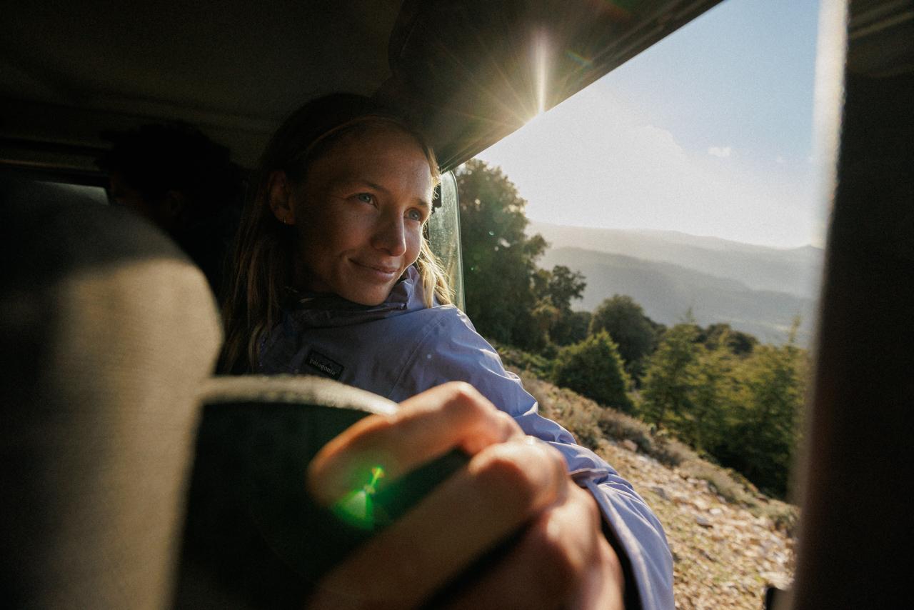 A woman in a blue jacket is sitting in the back of a vehicle, looking out at a scenic landscape.