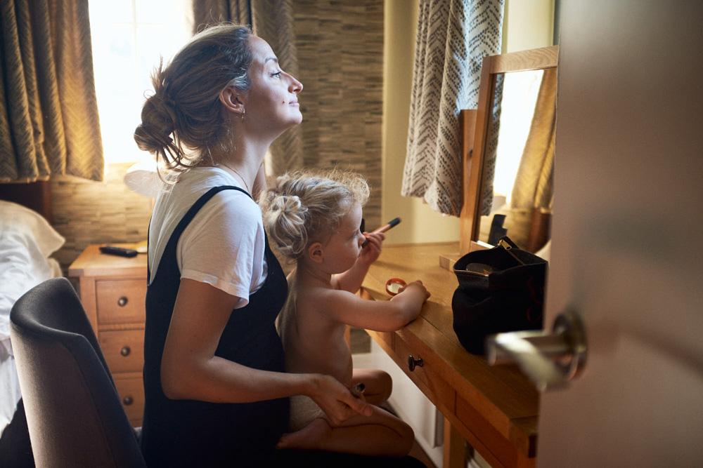 A woman sits at a desk, holding a toddler on her lap as she applies makeup. The woman has her eyes closed, and the child is playing with a makeup brush.