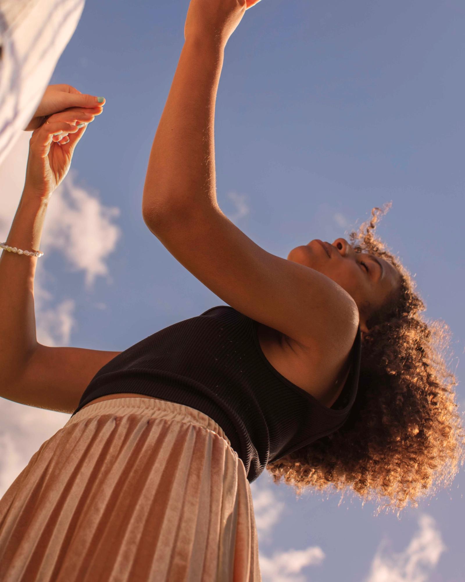 A woman with curly hair and a black tank top is reaching up towards something out of frame, with her arms stretched up and her face tilted up towards the sky.