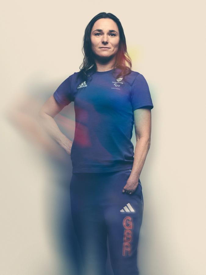 A woman with long dark hair wearing a blue Adidas t-shirt and matching blue Adidas tracksuit pants, posing with her left hand in her left pocket.