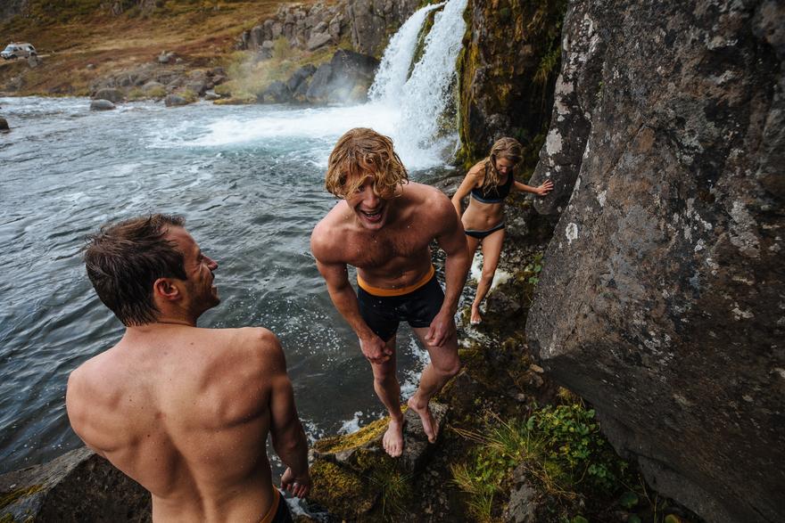 Two men and a woman are standing on a rocky outcrop near a waterfall, with the woman wearing a black bikini and the men wearing black swim shorts.