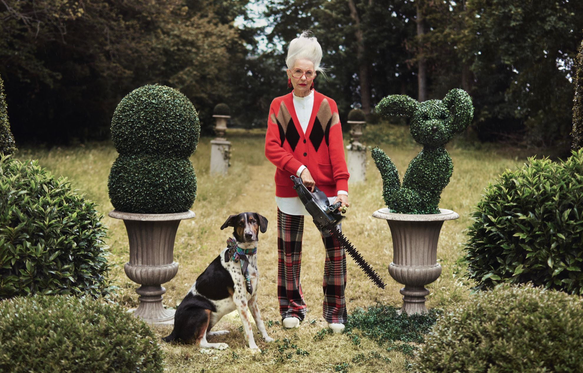 An elderly woman stands in a garden holding a hedge trimmer, surrounded by topiary animals and plants.