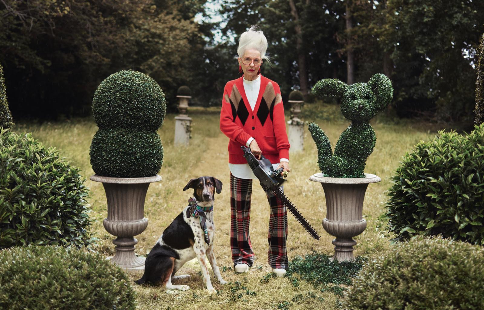 An elderly woman stands in a garden holding a hedge trimmer, surrounded by topiary animals and plants.