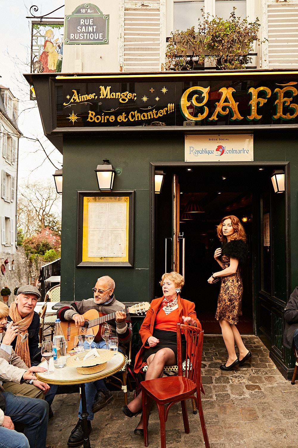 A group of older adults, some with guitars, sitting at a cafe in Paris, with a woman in a red coat and a man in a grey sweater playing a guitar, while others watch and drink wine.
