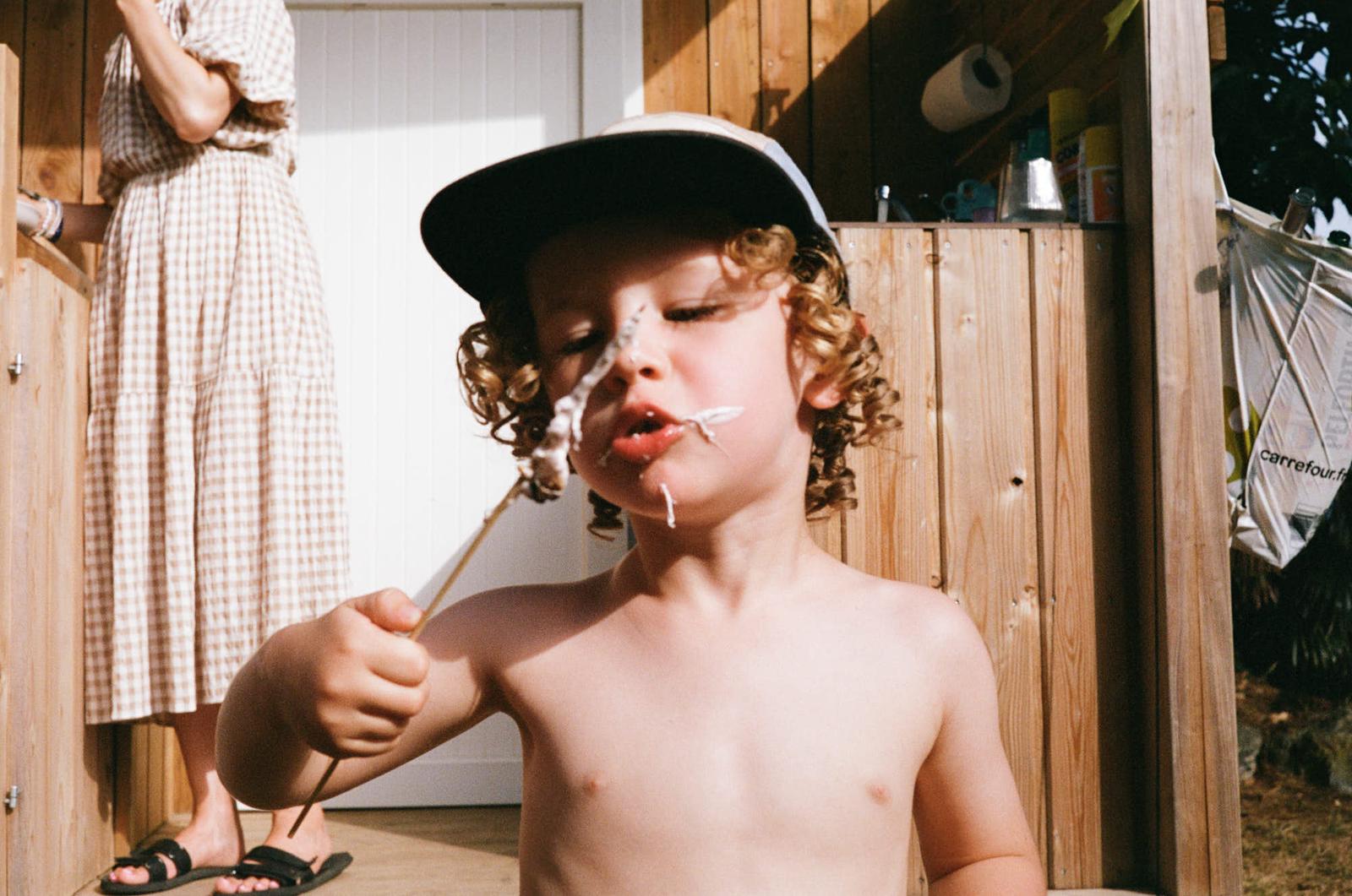 A young boy with curly hair and a baseball cap, covered in whipped cream, looks directly at the camera with a mischievous expression. He is shirtless and holding a spoon in his.