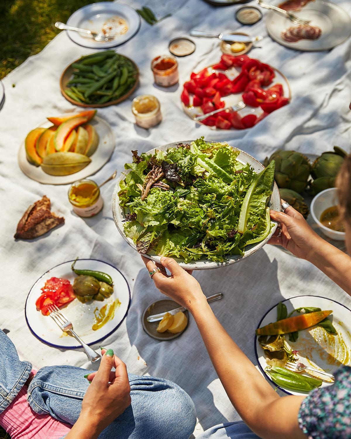 A woman holding a large bowl of mixed greens salad, surrounded by various food items and plates on a white tablecloth in a park or outdoor setting.