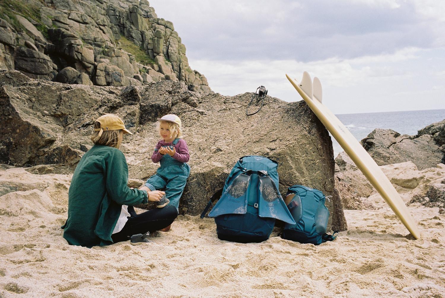A woman in a green jacket and a child in blue overalls are sitting on a beach, surrounded by surfboards and beach equipment, with a rocky cliff in the background.