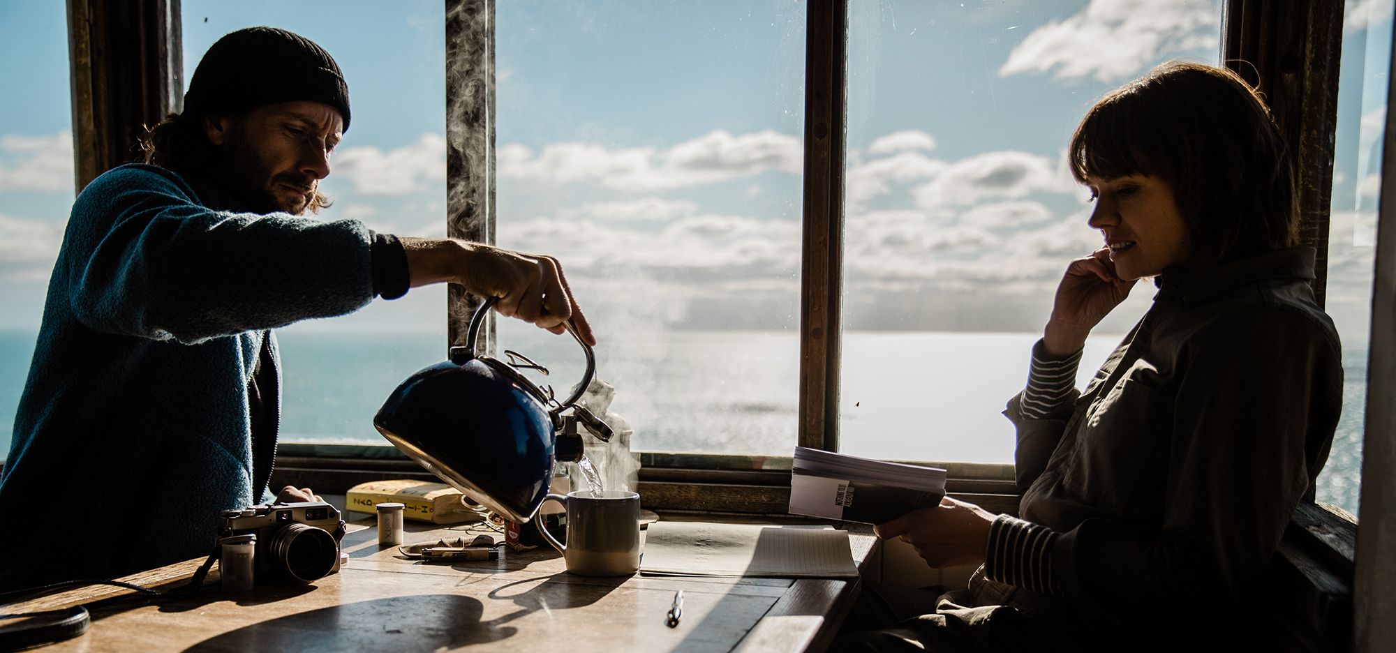 A man and a woman sit at a table by a window, gazing out at the ocean. The man is pouring a kettle of steamy water into a cup, while the woman sits with her hand to her chin,.