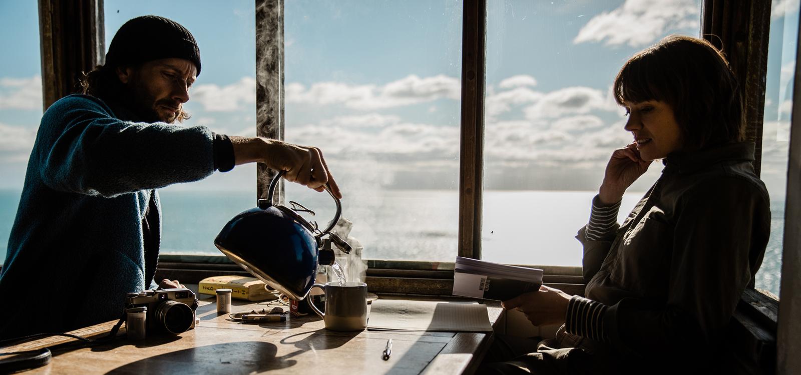 A man and a woman sit at a table by a window, gazing out at the ocean. The man is pouring a kettle of steamy water into a cup, while the woman sits with her hand to her chin,.