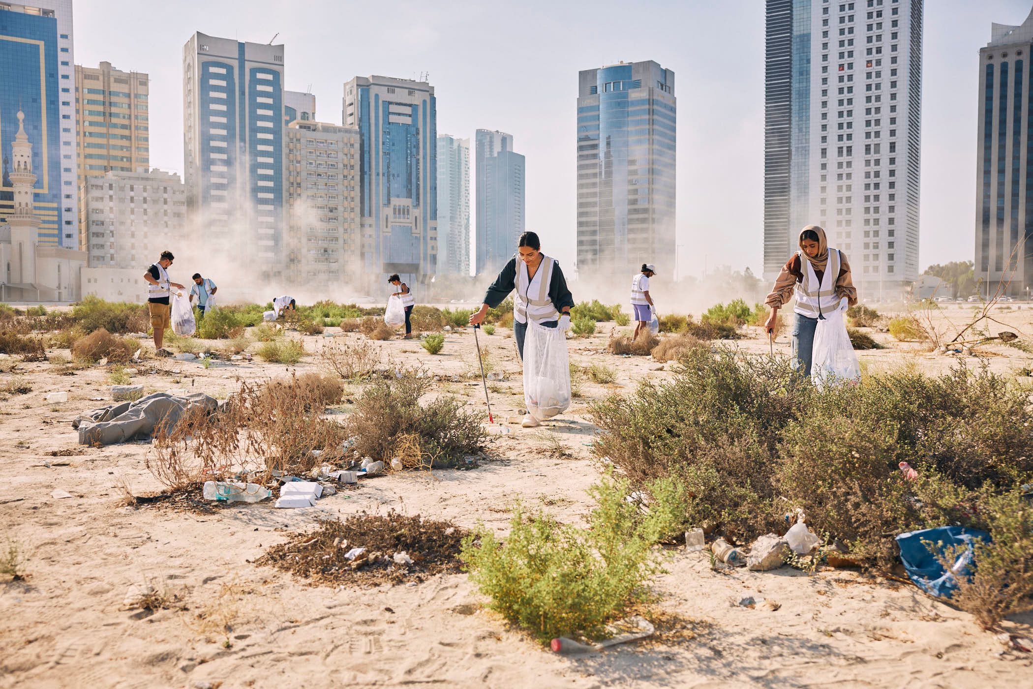 A group of people in white vests and trash bags walk through a dry, sandy area with scrubby bushes, collecting trash. In the background, a cityscape of tall, modern buildings.