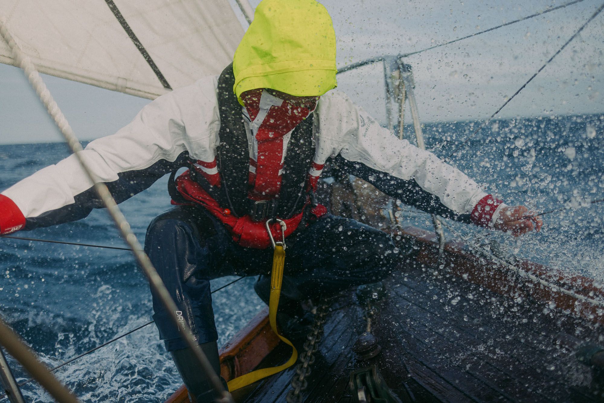 A person in a yellow rain hat, red and white life jacket, and blue wetsuit-like pants, is crouing on a boat in the middle of the ocean, with a white sail in the background.