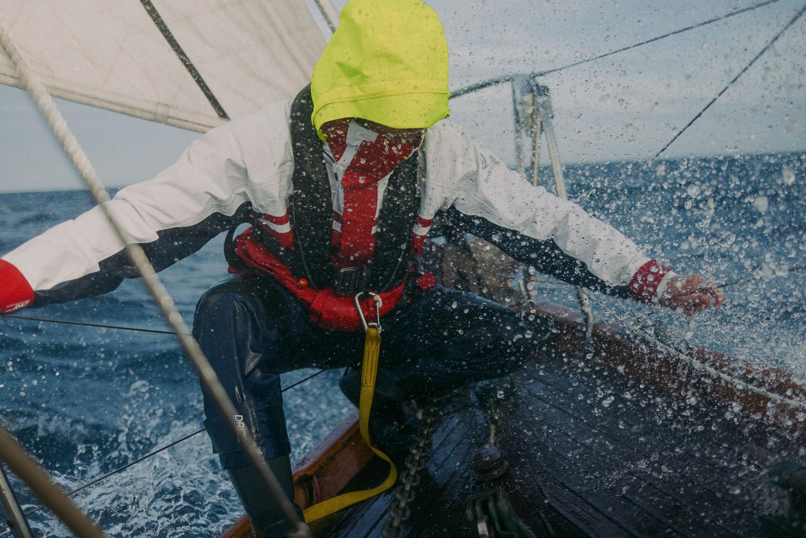 A person in a yellow rain hat, red and white life jacket, and blue wetsuit-like pants, is crouing on a boat in the middle of the ocean, with a white sail in the background.