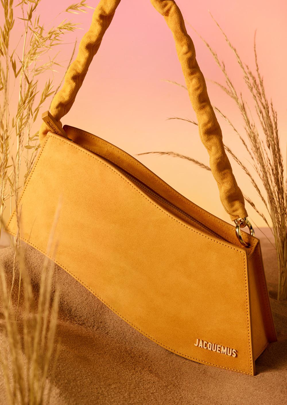 A handbag made of yellow suede leather, with a long strap and a gold clasp, sitting on a sandy beach with some tall grasses in the background.