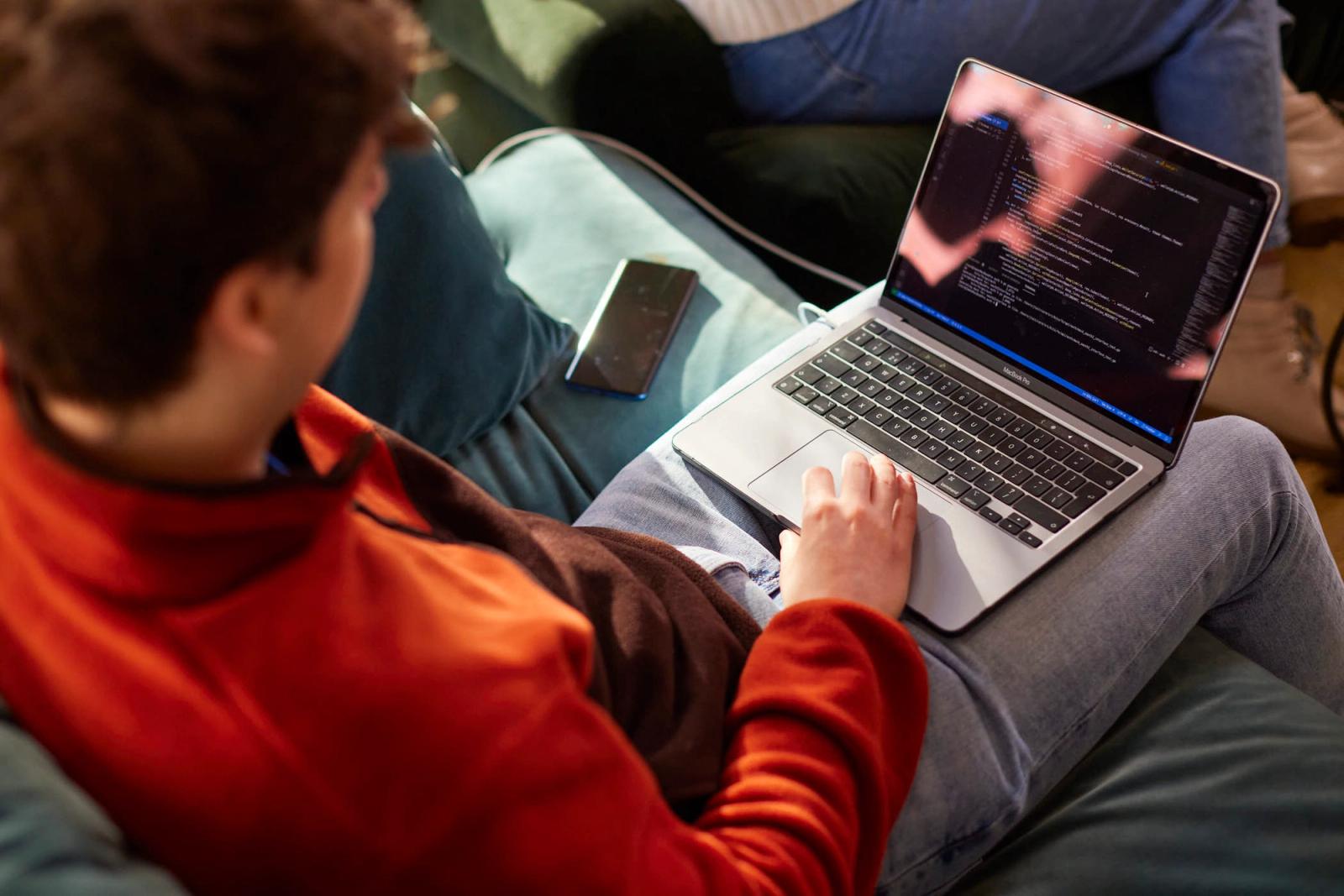 A person in a red sweater sits on a couch, looking at a laptop screen. The screen displays a code editor with a dark background and white text. The person's face is not visible,.