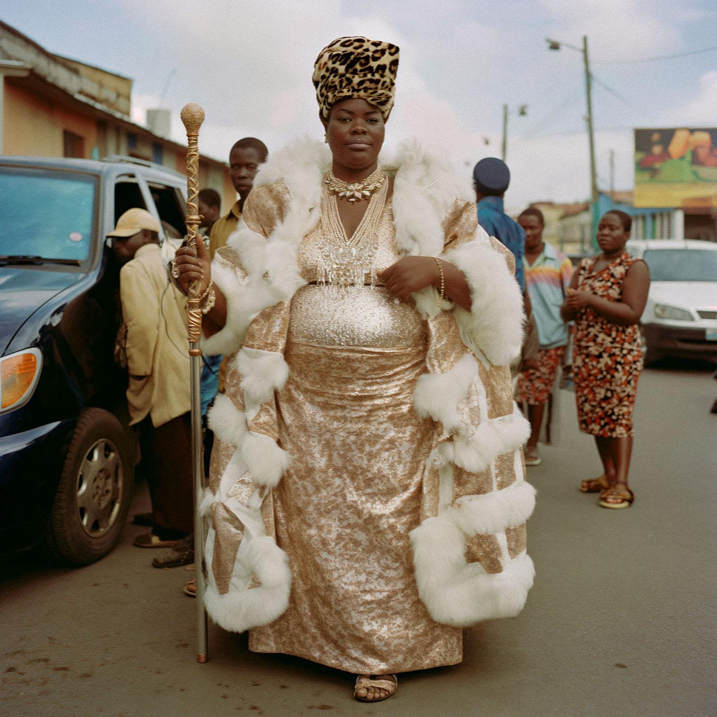 A woman in a gold and white fur-trimmed coat, leopard-print headscarf, and gold jewelry stands in a city street, holding a gold-tipped staff.