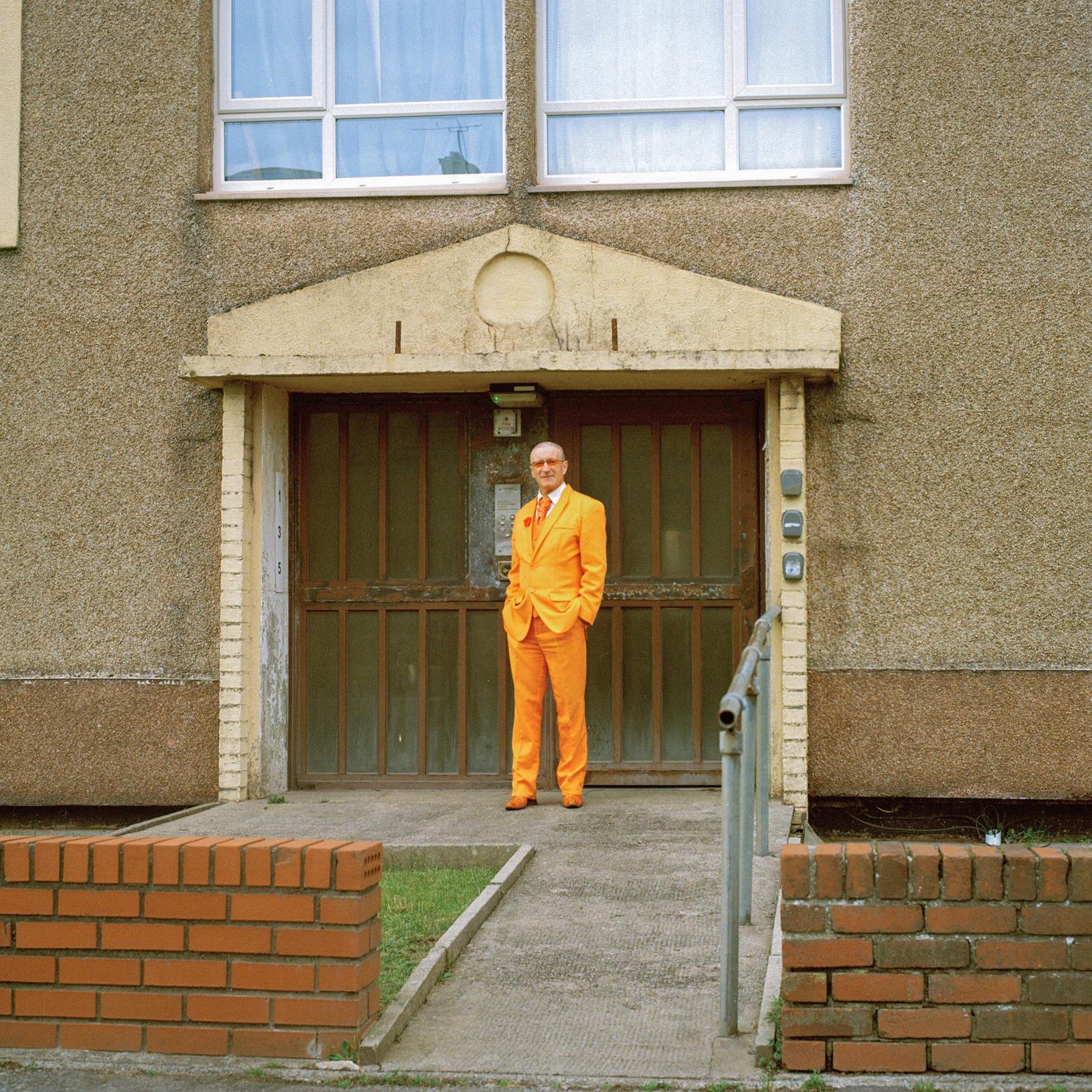 A man in a bright orange suit stands in front of a grey concrete building with a large metal door.