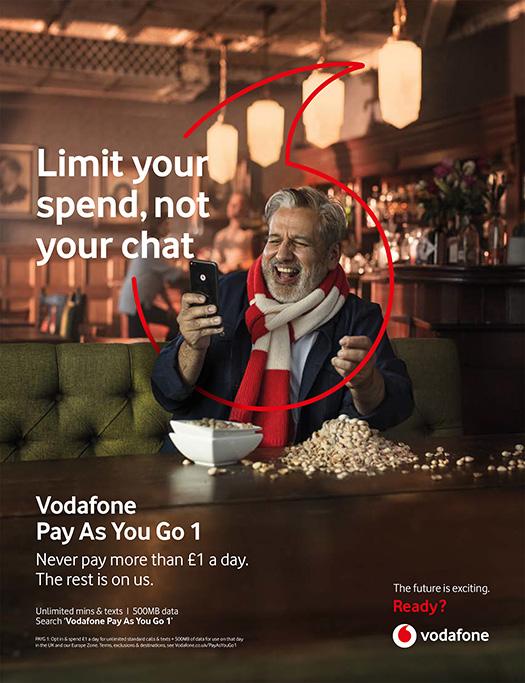 A man sitting in a restaurant, holding a phone and laughing, with a bowl of food in front of him.