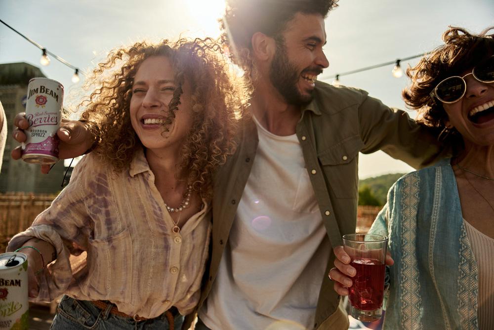 Three people, two women and one man, laughing and enjoying Jim Beam Red Stewing Berries Bourbon Spritz on a rooftop or outdoor location with string lights in the background.