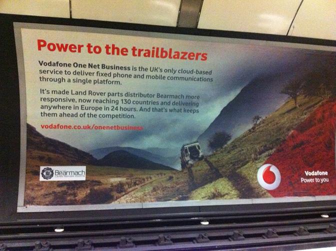 A large billboard in a subway station featuring a Vodafone ad with a scenic landscape of a mountain road with a car driving on it, with a Vodafone logo in the top left corner.