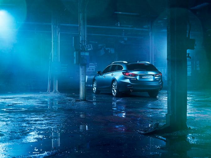 A silver Mazda car is parked in a dimly lit, abandoned warehouse with a few scattered boxes and crates in the background.