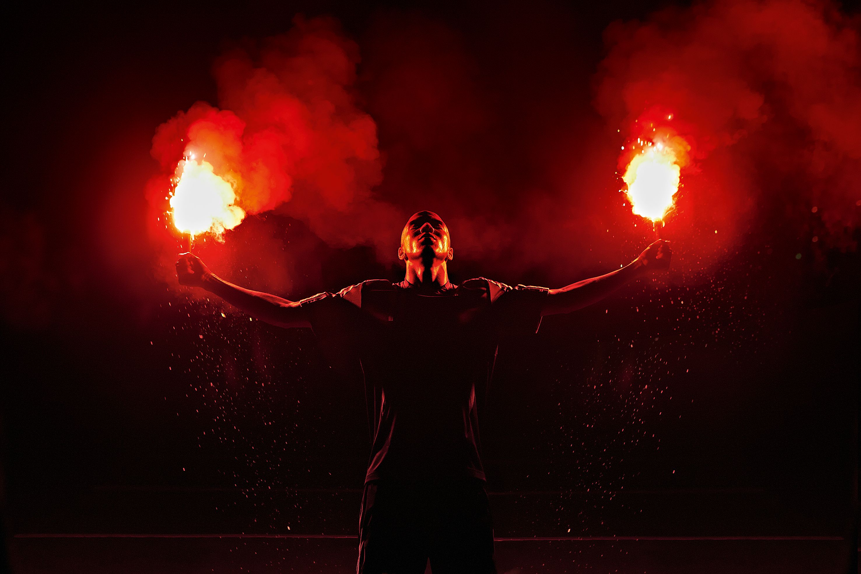 A man in a black shirt and pants is standing in a dark room, surrounded by flames, with his arms outstretched and his face illuminated by the fire.