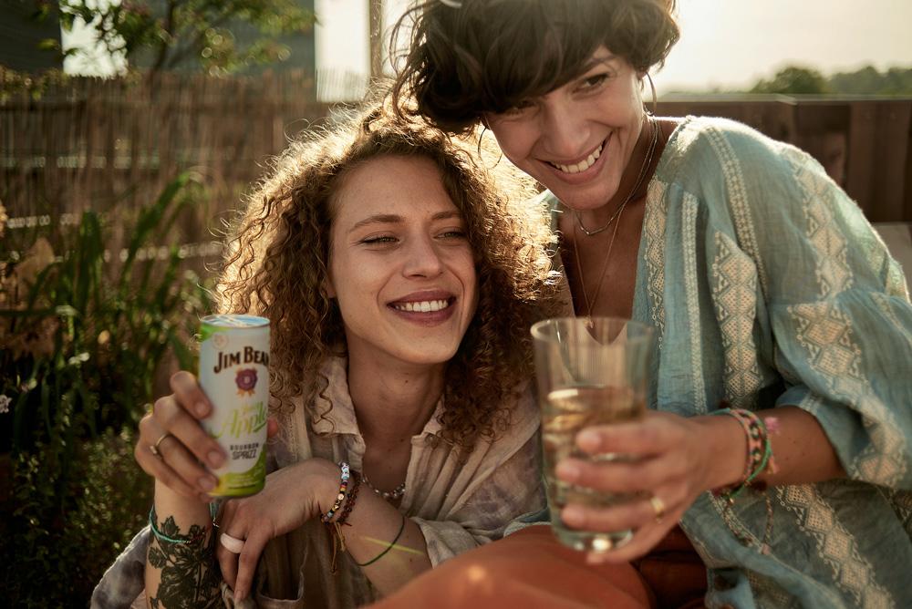 Two women laughing and hugging, one holding a Jim Beam can, outdoors by a wooden fence.