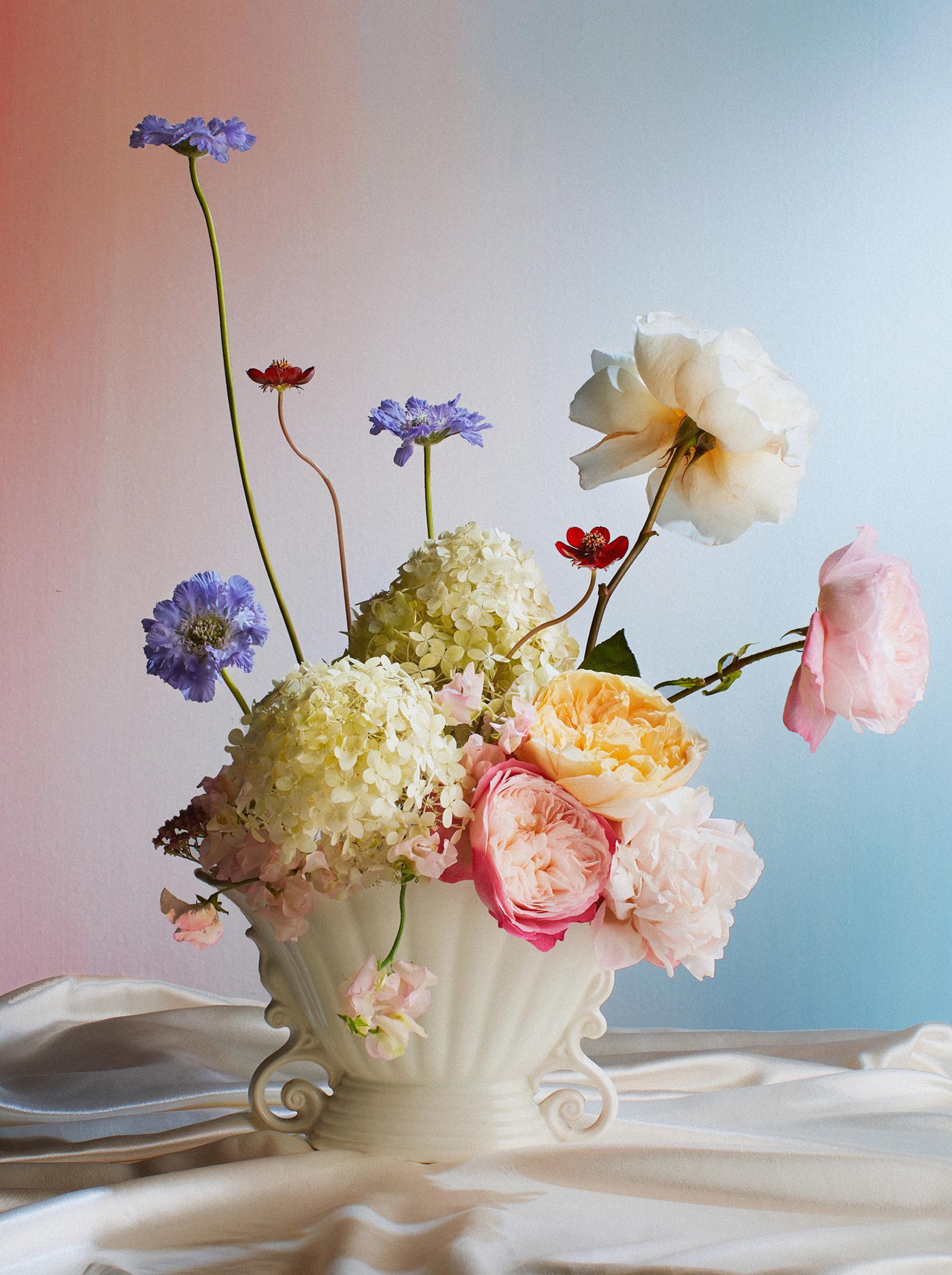 A bouquet of flowers in a white vase, featuring a mix of flowers including roses, hydrangeas, and wildflowers, arranged in a white ceramic vase on a table with a white tablecloth.