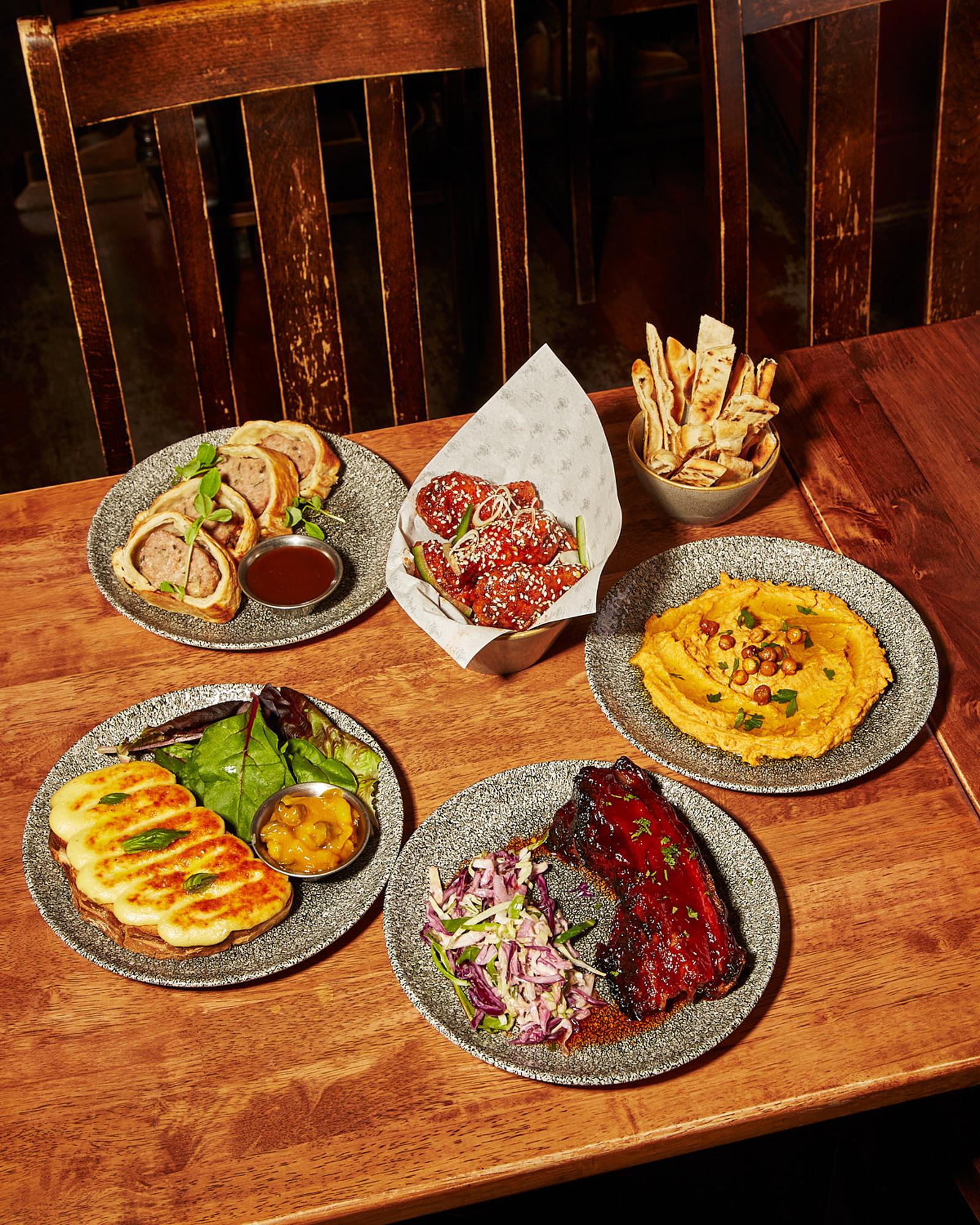 A selection of various dishes, including a plate of ribs, a bowl of hummus, and a plate of breadsticks, on a wooden table in a restaurant or cafe.