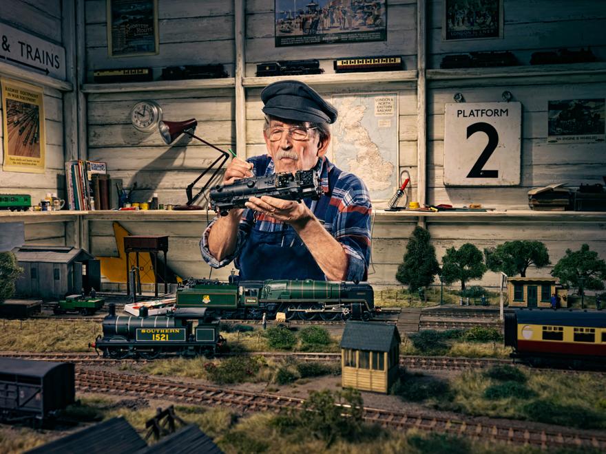 An elderly man in a blue and red striped shirt, wearing a black hat and glasses, is sitting in front of a model train set, carefully adjusting a train's engine.