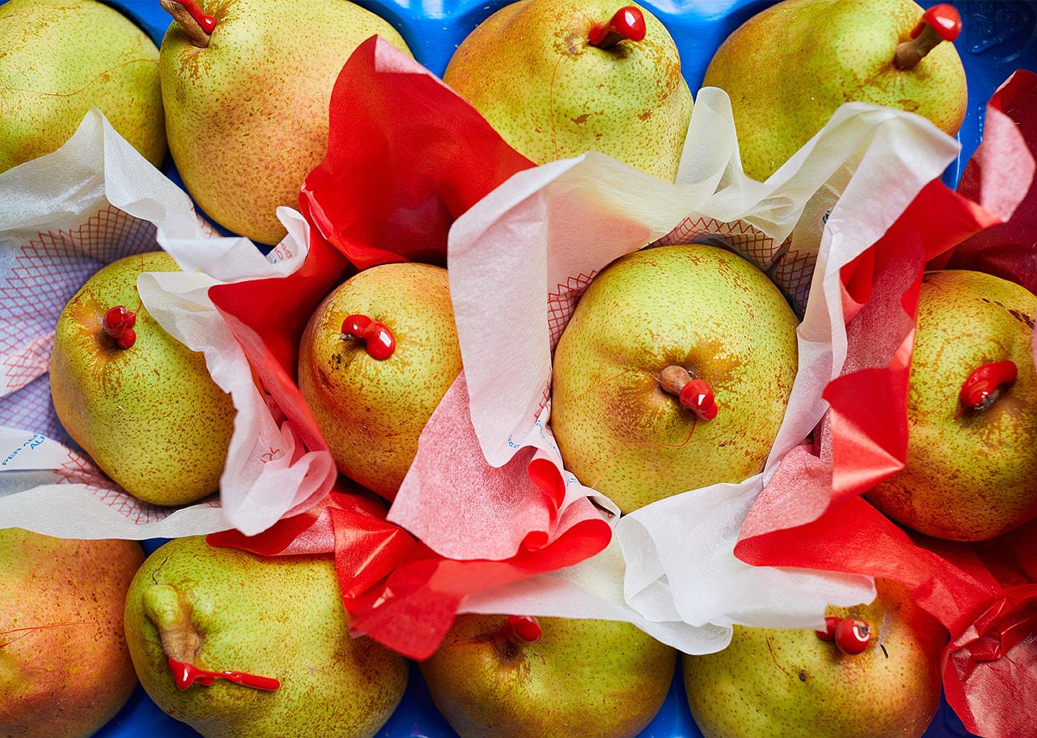 A still life featuring a large blue ceramic bowl containing various fresh green and red pears, some wrapped in paper.