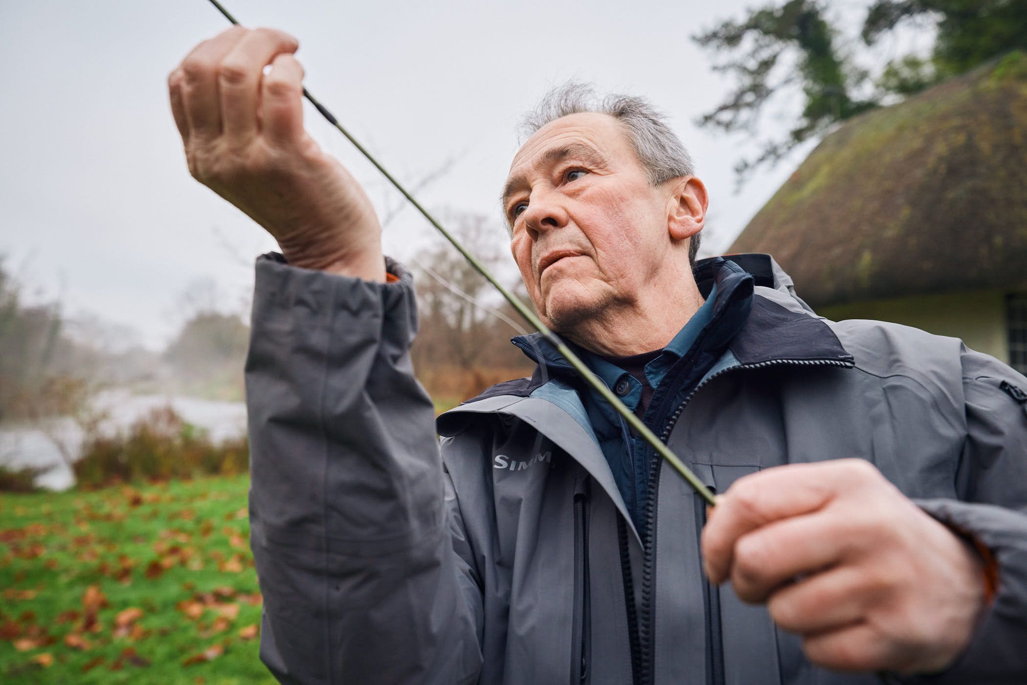 An older man in a gray jacket holds a fishing rod in front of a small cottage. The man's face is serious, and he appears to be focused on his fishing.