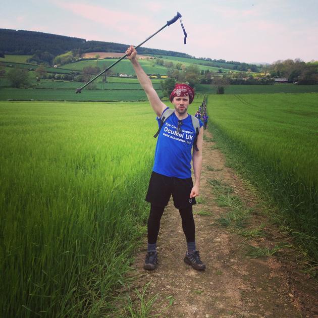 A person in a blue t-shirt and black leggings walking on a dirt path in a green field, holding a walking pole in the air.