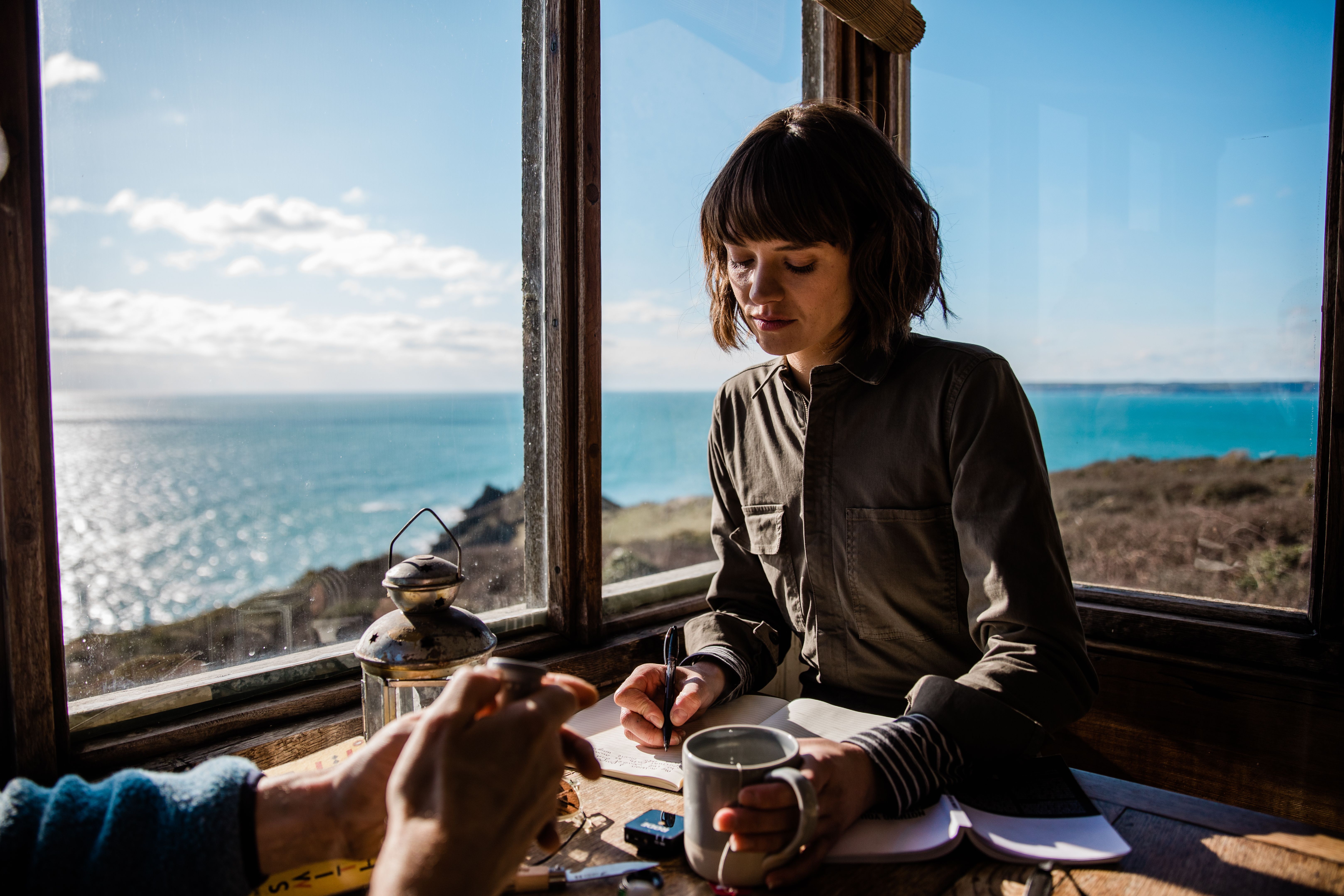 A woman sits at a table by a window, writing in a notebook while looking out at the ocean.