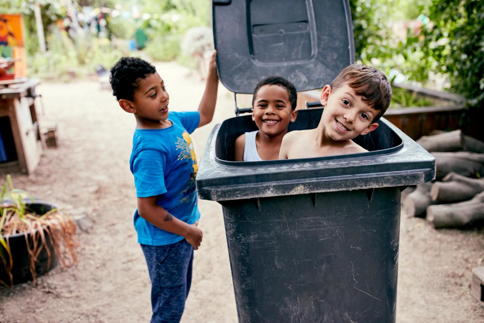 Three children peeking out of a large trash bin, grinning mischievously. They appear to be having fun and are likely playing a game or playing pranks.